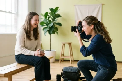 A female photographer kneels down to take a photo of a young woman laughing during a photoshoot in a natural light studio. This image demonstrates the hands-on work and client interaction involved in running a photography company, a key consideration for anyone researching how to start a photography business.