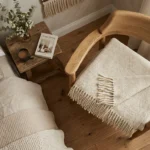 A cozy bedroom corner with a natural wood chair holding a folded wool blanket with fringes. Next to it is a bed and a rustic side table with a mug and a book titled 'Cozy Living', perfectly illustrating the comfort of wool that needs careful maintenance, often searched for by those looking for how to wash wool blanket.