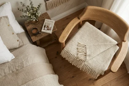 A cozy bedroom corner with a natural wood chair holding a folded wool blanket with fringes. Next to it is a bed and a rustic side table with a mug and a book titled 'Cozy Living', perfectly illustrating the comfort of wool that needs careful maintenance, often searched for by those looking for how to wash wool blanket.