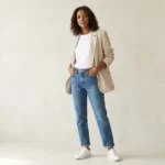 A full-body photograph of a woman with shoulder-length curly hair, wearing a beige unstructured linen blazer over a white t-shirt and light-wash straight-leg jeans with white sneakers. She is standing with her hands in her pockets and looking confidently at the camera against a textured beige studio background with soft window light. Trying to explain what is Blazertje.