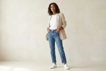 A full-body photograph of a woman with shoulder-length curly hair, wearing a beige unstructured linen blazer over a white t-shirt and light-wash straight-leg jeans with white sneakers. She is standing with her hands in her pockets and looking confidently at the camera against a textured beige studio background with soft window light. Trying to explain what is Blazertje.