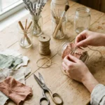 Flat-lay of a home craft workspace with upcycled glass jars, natural twine, and fabric scraps, with hands shaping materials in soft natural light.